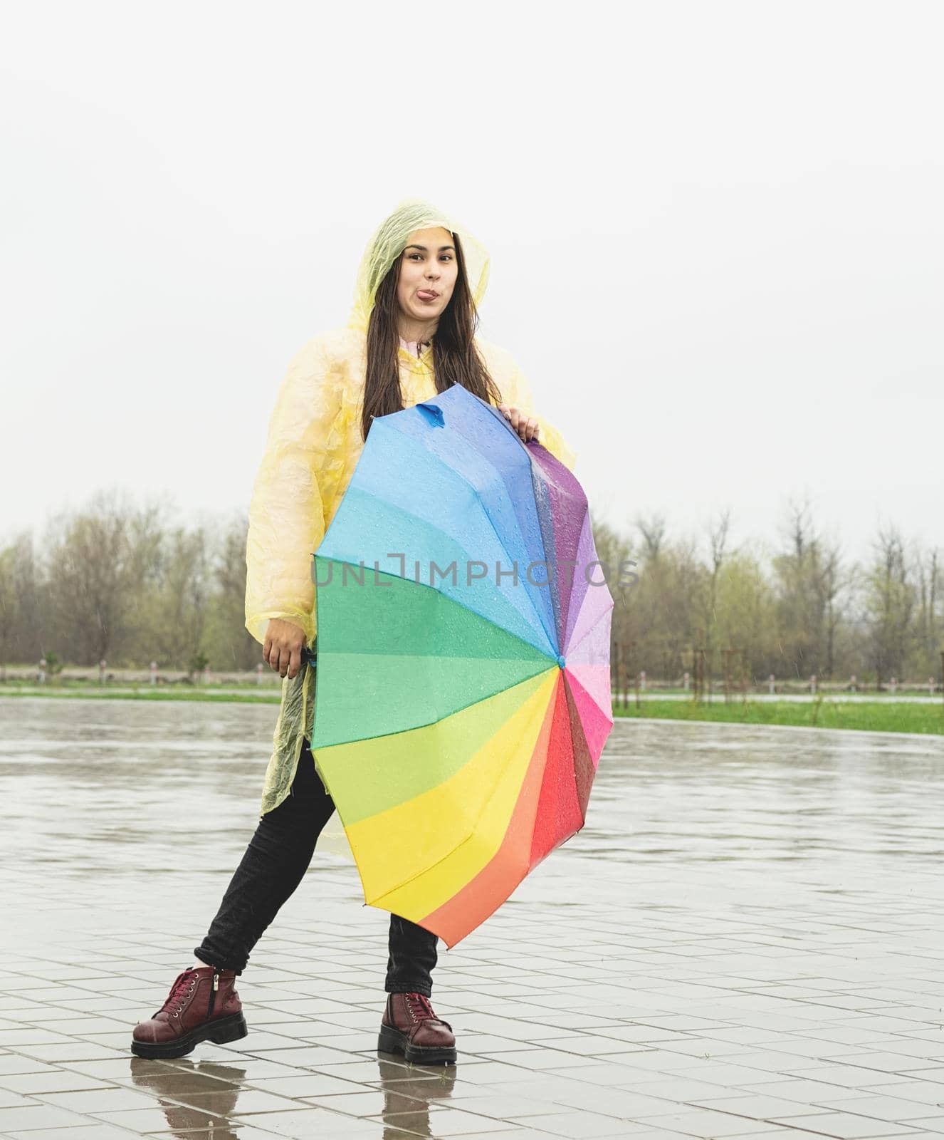 Beautiful brunette woman in yellow raincoat holding rainbow umbrella out in the rain, making funny face showing tongue out