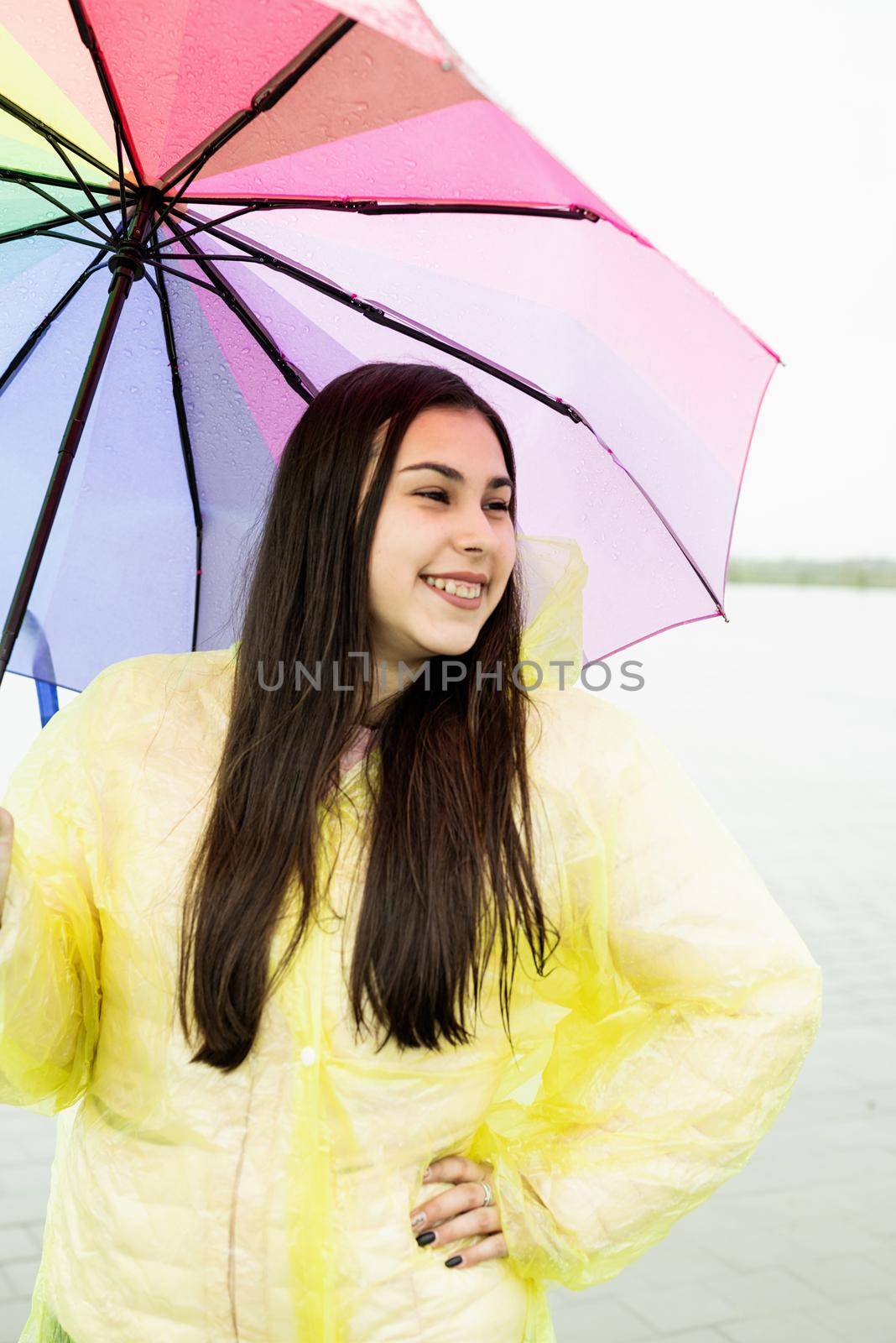 Beautiful smiling brunette woman in yellow raincoat holding rainbow umbrella out in the rain