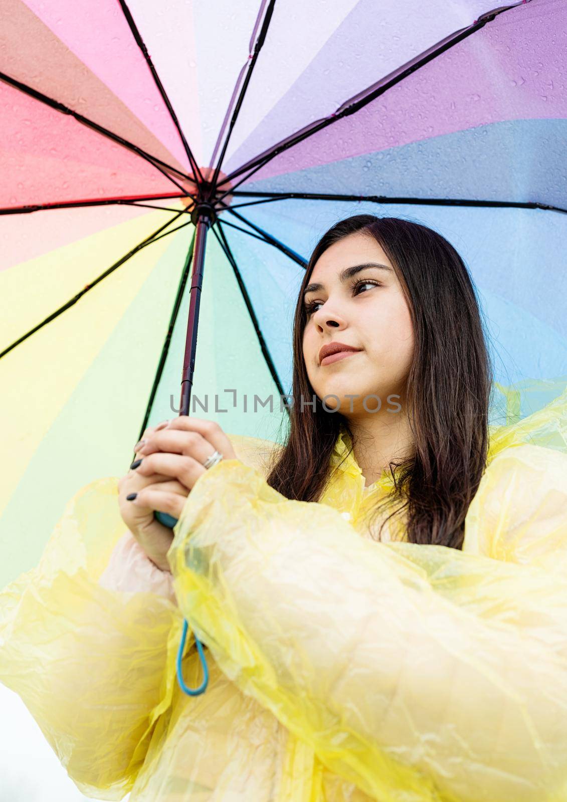 Beautiful smiling brunette woman in yellow raincoat holding rainbow umbrella out in the rain