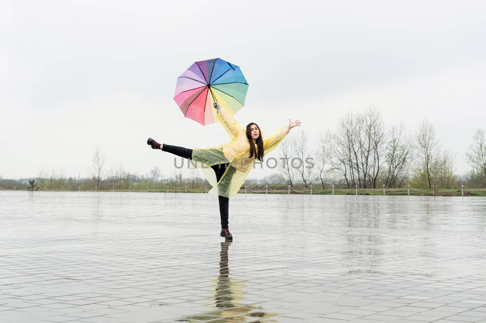 Beautiful brunette woman in yellow raincoat holding rainbow umbrella out in the rain
