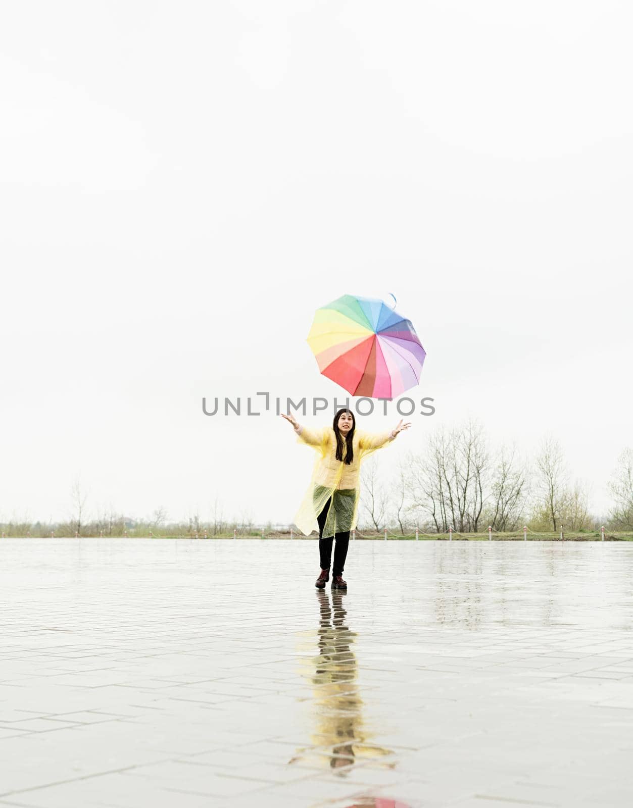 Beautiful brunette woman with funny face in yellow raincoat catching colorful umbrella outdoors in the rain