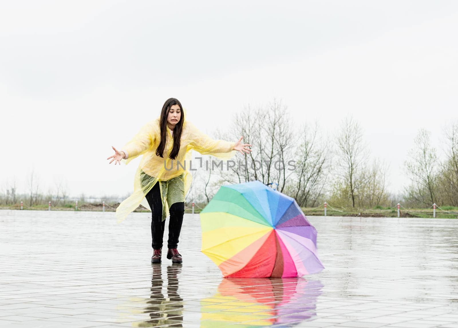 Beautiful brunette woman with funny face in yellow raincoat catching colorful umbrella outdoors in the rain. Rainbow umbrella lying on the ground