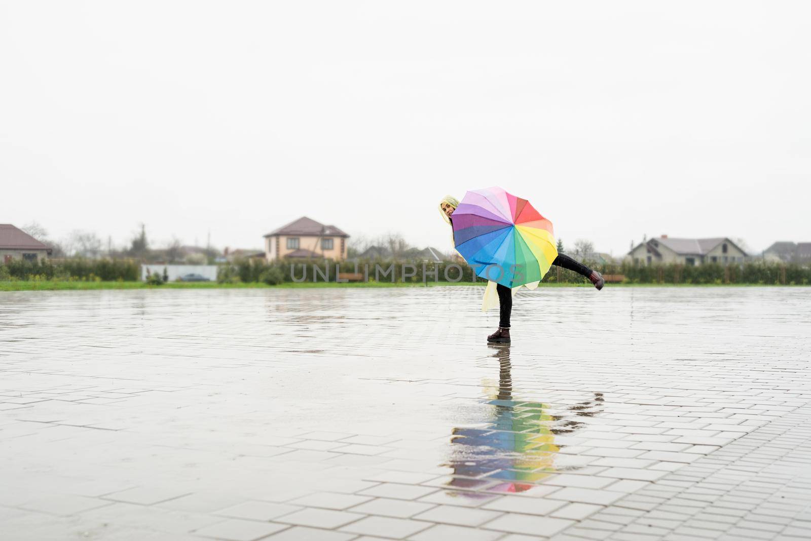 Beautiful brunette woman in yellow raincoat holding rainbow umbrella out in the rain