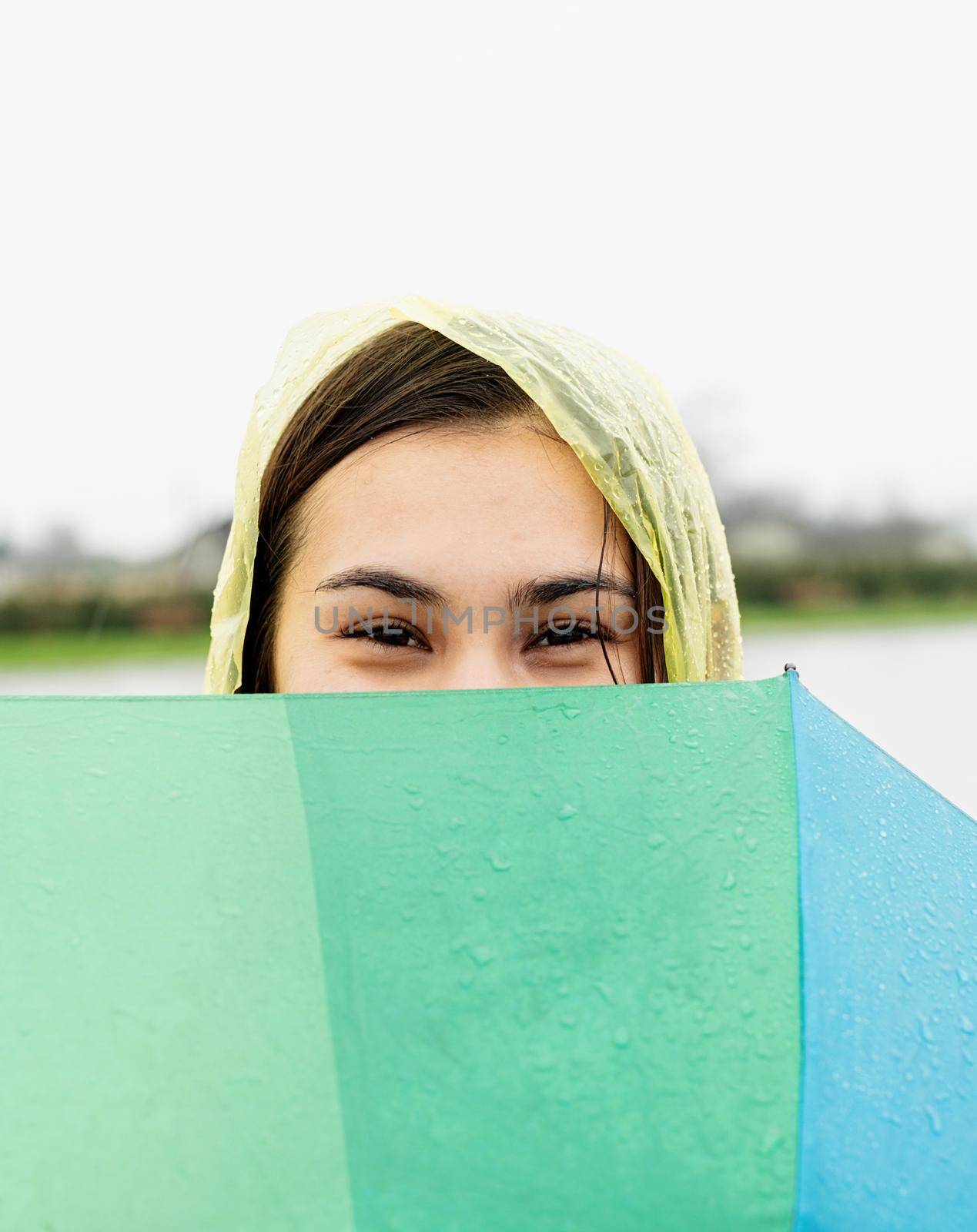 Beautiful smiling brunette woman in yellow raincoat holding rainbow umbrella out in the rain, covering her face with umbrella