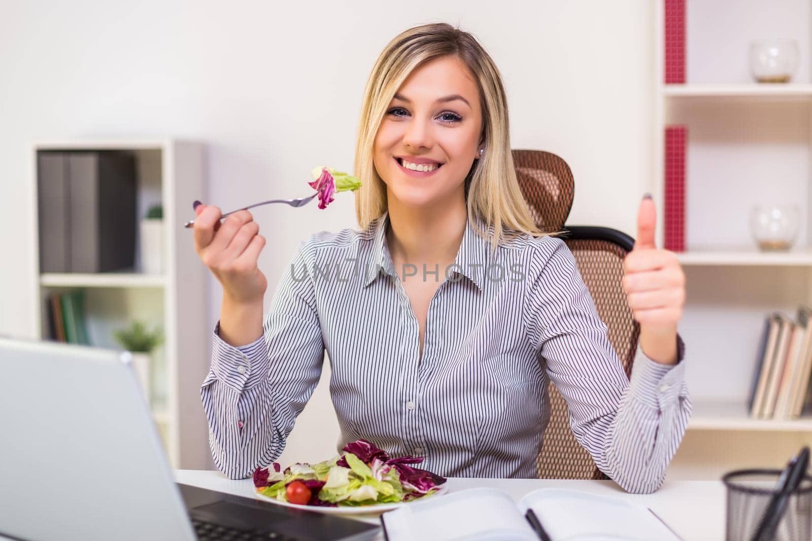Beautiful businesswoman enjoys eating salad and showing thumb up  while working in her office.