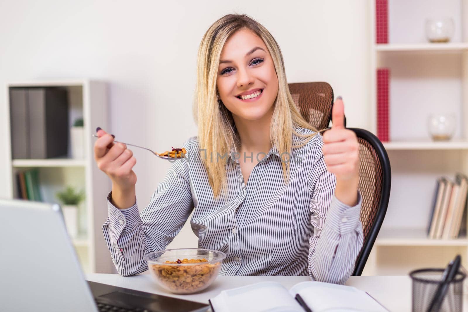 Beautiful businesswoman showing thumb up while eating corn flakes for breakfast and working in her office.