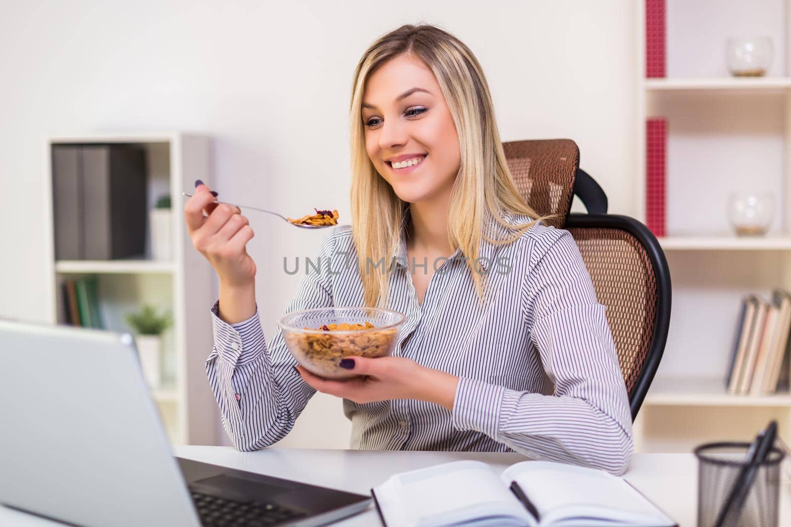 Beautiful businesswoman enjoys eating corn flakes for breakfast while working in her office.