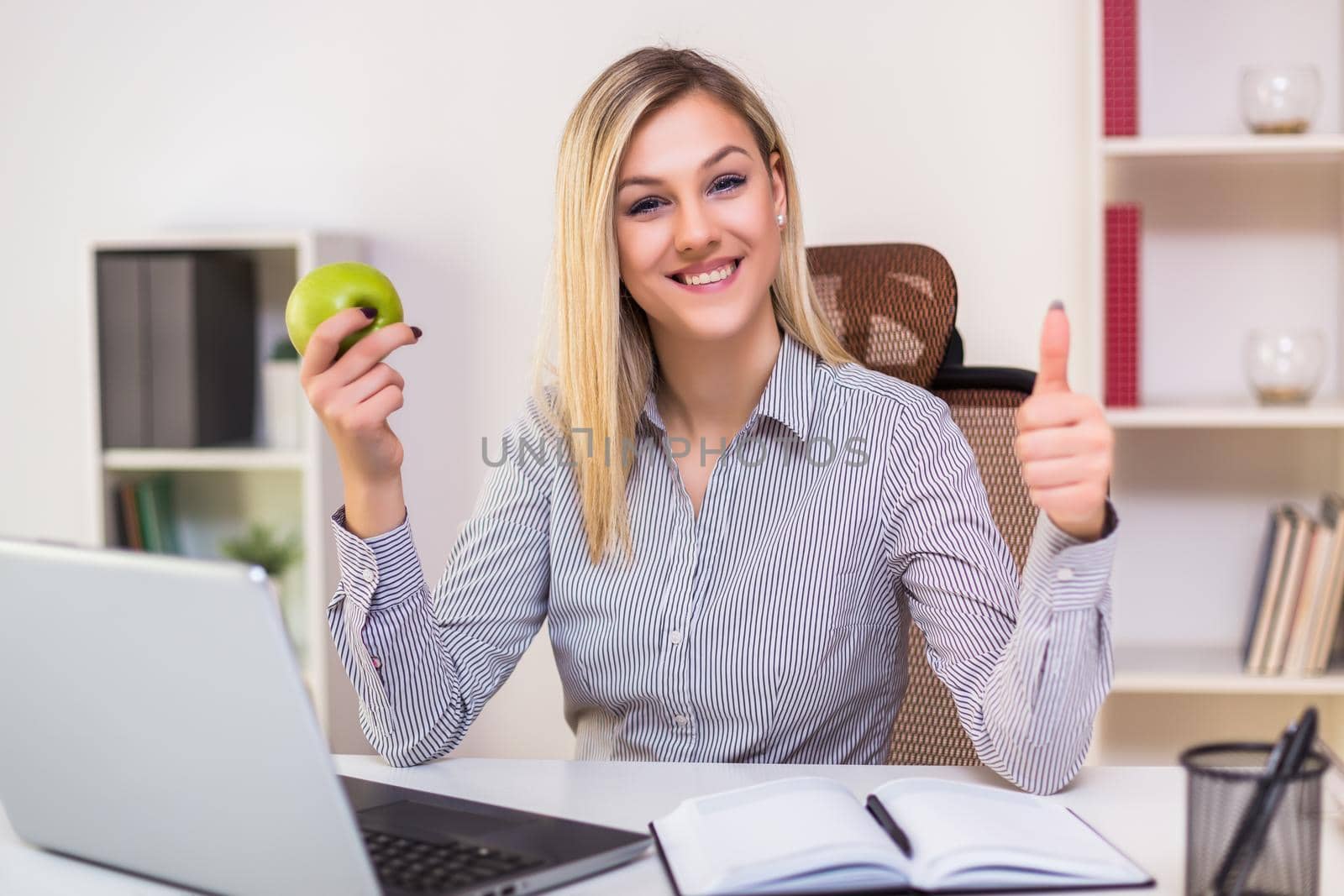 Businesswoman eating apple and showing thumb up while working in her office.