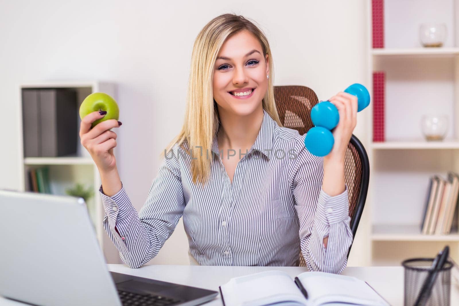 Beautiful businesswoman holding apple and weights while working in her office.