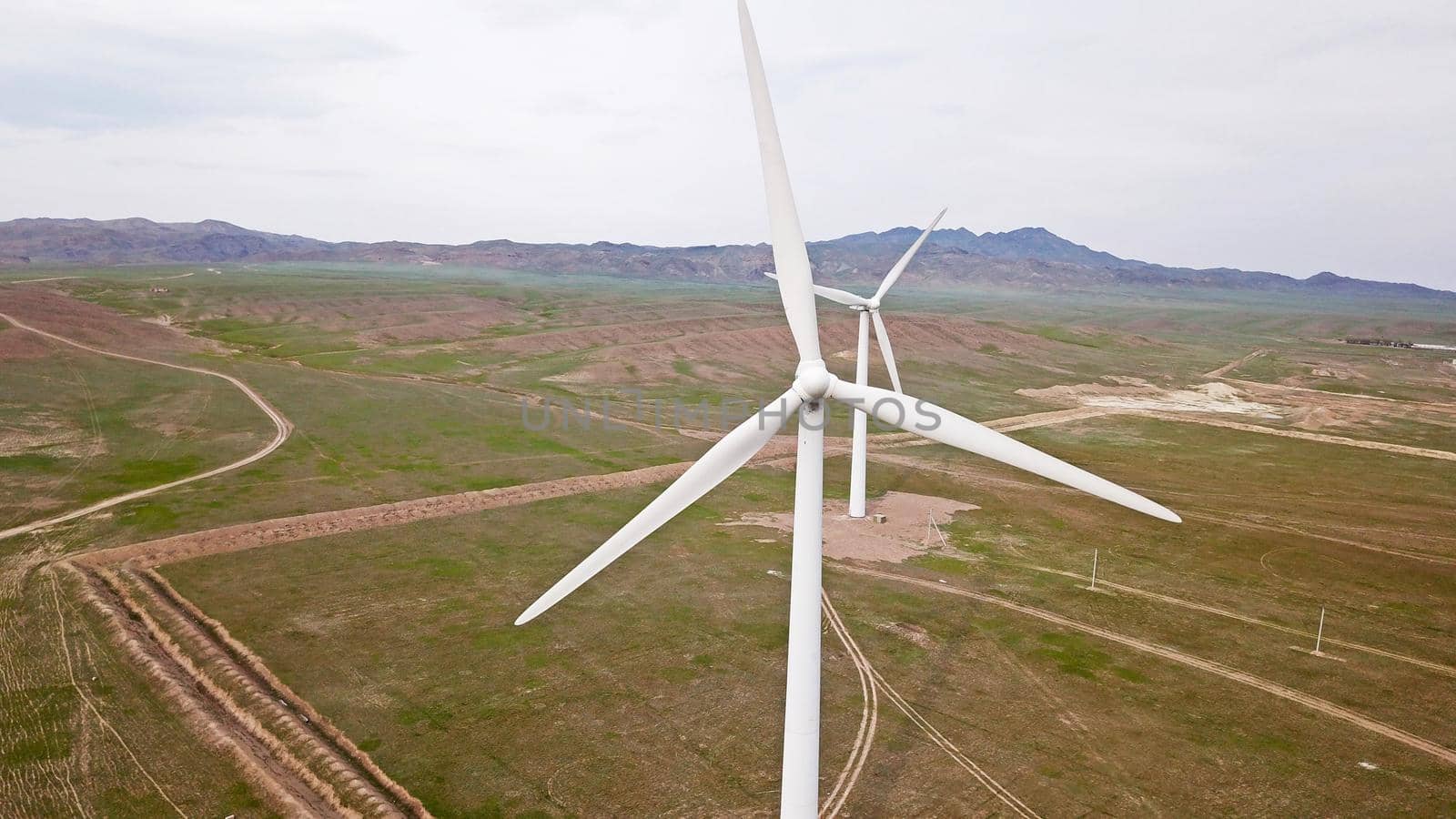 Windmills stand in the middle of green fields. Clean energy is generated. Cloudy weather, dark clouds. Green grass grows. High hills in the distance. Top view from drone. Industrial farm of Kazakhstan