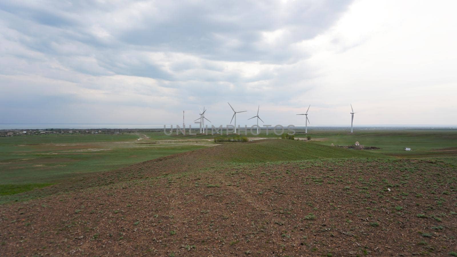 Windmills stand in the middle of green fields. Clean energy is generated. Cloudy weather, dark clouds. Green grass grows. High hills in the distance. Top view from drone. Industrial farm of Kazakhstan