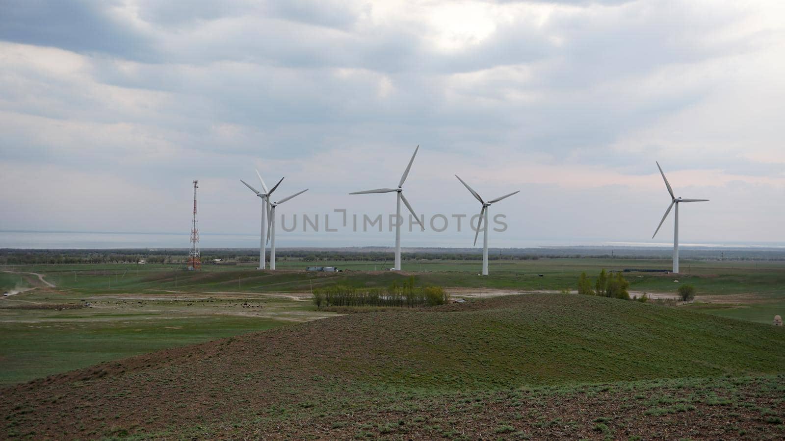 Windmills stand in the middle of green fields. Clean energy is generated. Cloudy weather, dark clouds. Green grass grows. High hills in the distance. Top view from drone. Industrial farm of Kazakhstan