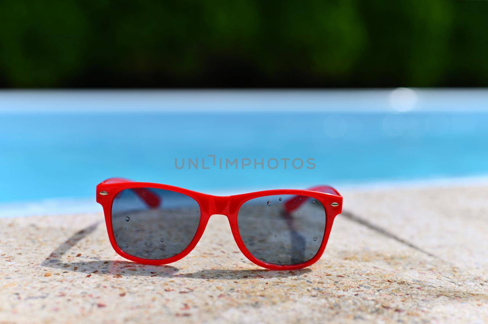 Summer background. Concept for summer and vacation. Red sunglasses by the pool in the background with blue water.