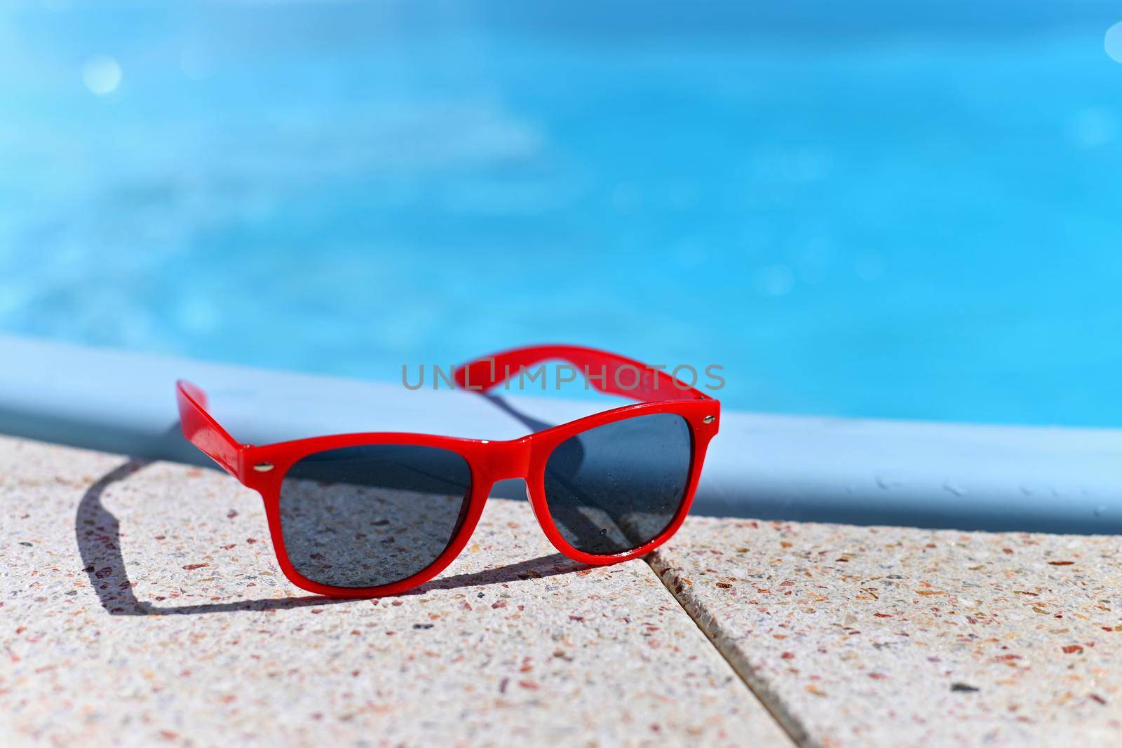 Summer background. Concept for summer and vacation. Red sunglasses by the pool in the background with blue water. by Montypeter