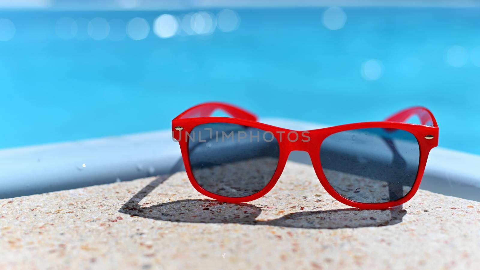 Summer background. Concept for summer and vacation. Red sunglasses by the pool in the background with blue water.