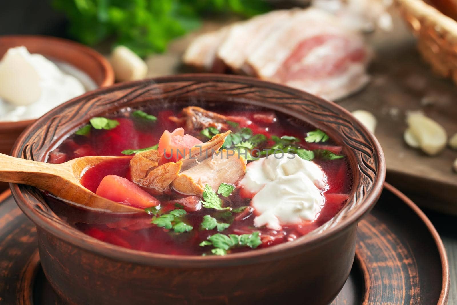 Freshly cooked borscht - traditional dish of Russian and Ukrainian cuisine in earthenware dishes with bacon, bread, sour cream and garlic, close up by galsand