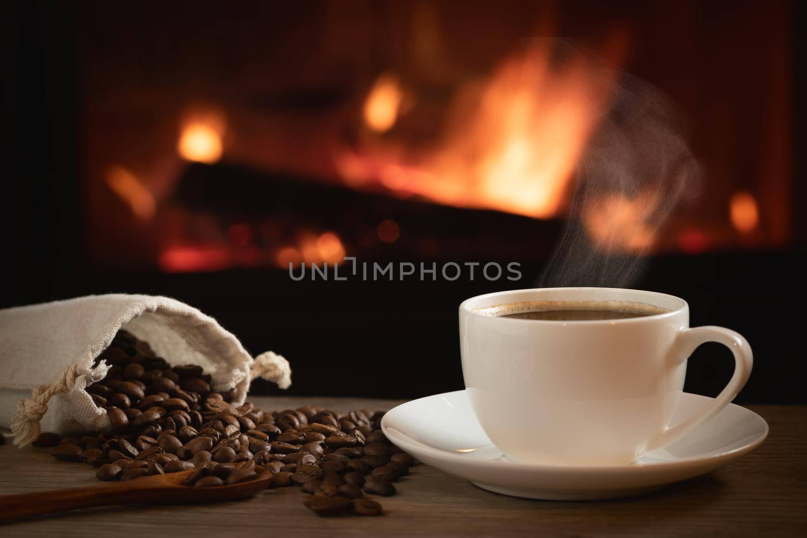 Cup of hot coffee and coffee beans in a bag on a wooden table in front of a burning fireplace. Selective focus.