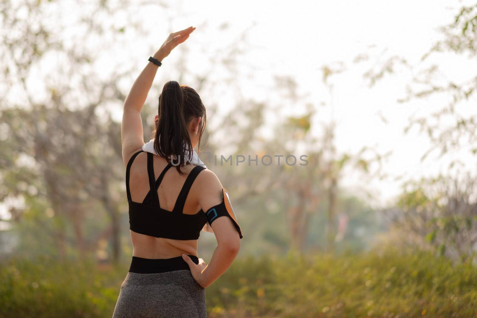 Athletic woman warming up before a workout standing facing the early morning rising sun at park.