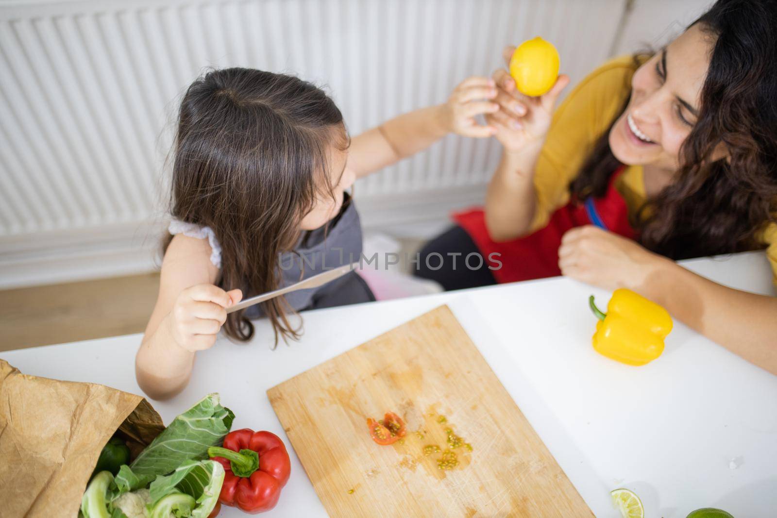 Happy little girl and her mother slicing lemons and tomatoes on cutting board. Smiling woman and young child at white talbe holding lemons. Daughter-mother cooking together