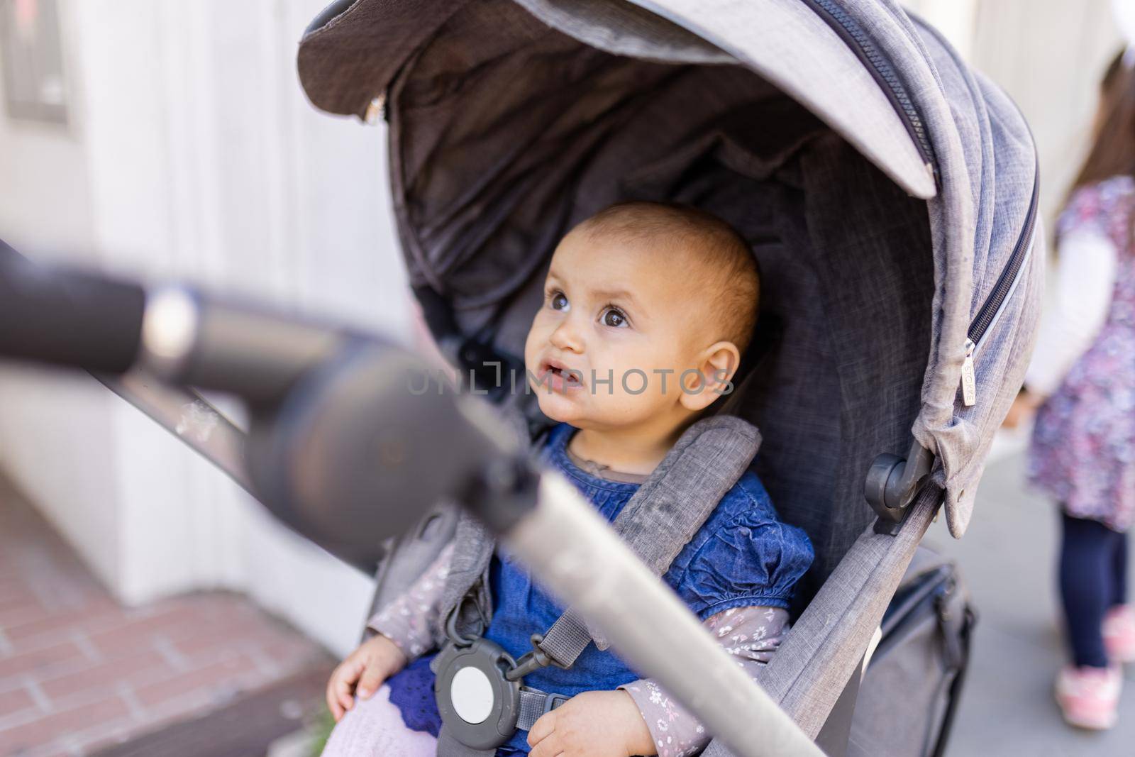 Adorable concerned-looking baby looking up in gray stroller on the sidewalk. Portrait of cute infant in blue dress resting in stroller in the street. Peaceful toddlers outing