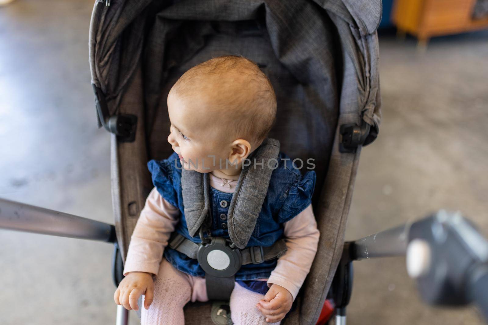 Adorable view of happy baby looking away in gray stroller with blurry background. Portrait of cute infant in blue dress resting in stroller inside a restaurant. Peaceful toddlers indoors
