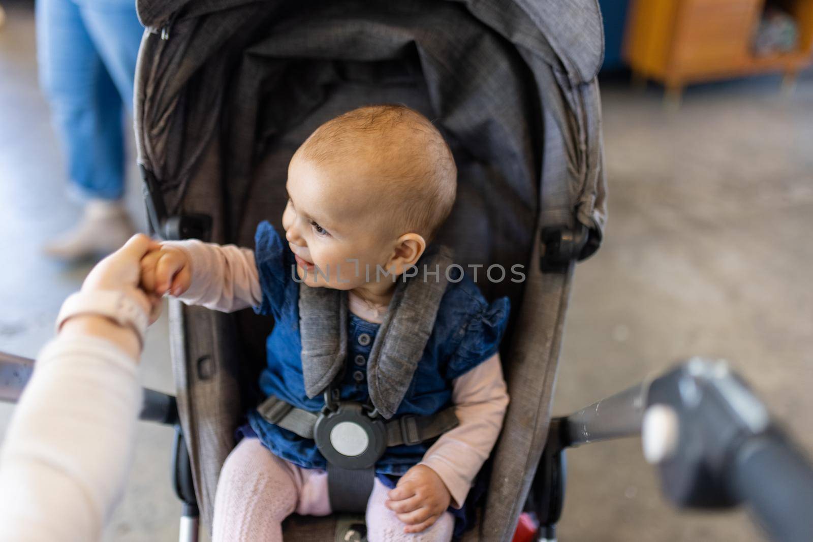 Adorable view of happy baby holding the hand of her sister in gray stroller with blurry background. Portrait of cute infant in blue dress resting inside a restaurant. Peaceful toddlers indoors