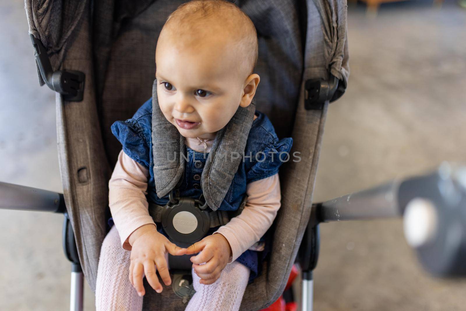 Adorable view of happy baby in gray stroller with blurry background. Portrait of cute infant in blue dress resting in stroller inside a restaurant. Peaceful toddlers indoors