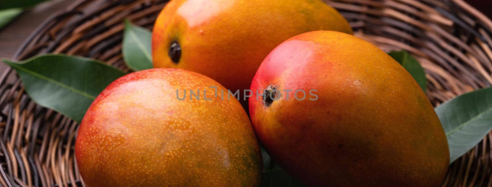 Mango. Close up of fresh ripe mango fruit on a bamboo sieve over dark wooden table background with green leaves.
