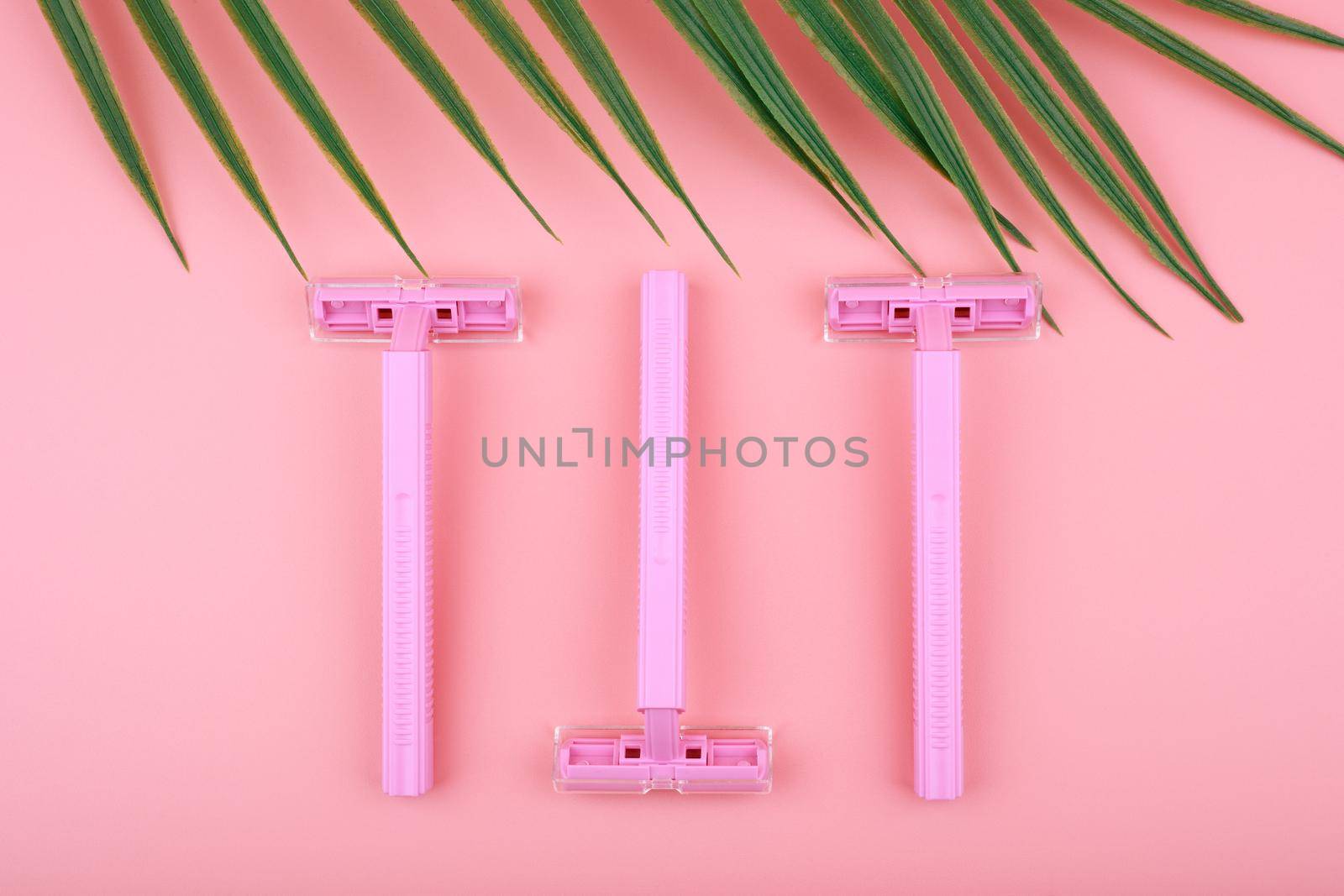 Top view of three pink razors against pink background with palm leaf. Minimal concept of shaving and epilation