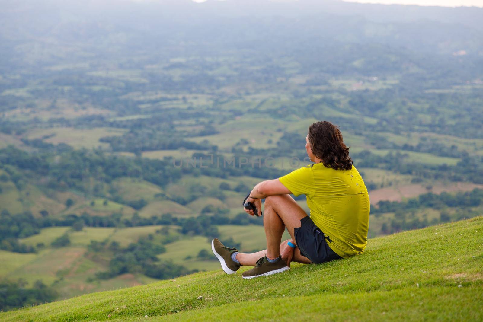 In summer, a man with long hair sits on the green slope of the mountain, looking into the distance at the valley. Dominican Republic, sunset in the mountains