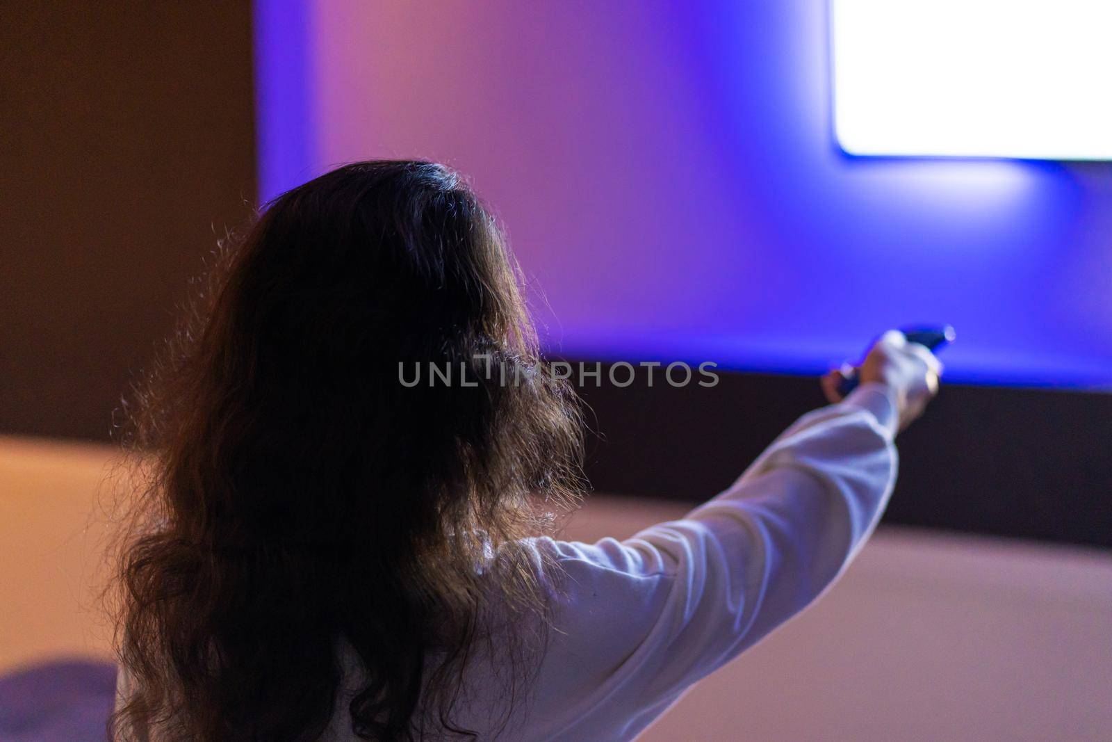 A person watches TV with a remote control in hand, a view from the back.