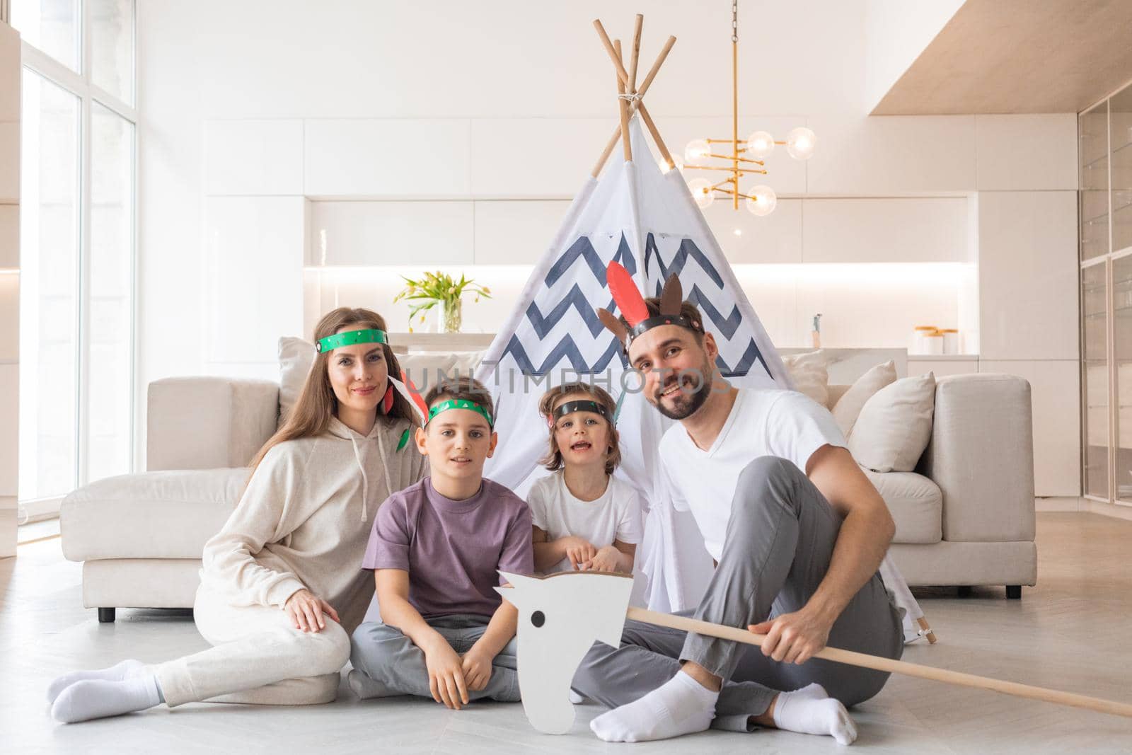 Happy family of parents and two children playing indian at home, wigwam tent, toy horse, feather roach