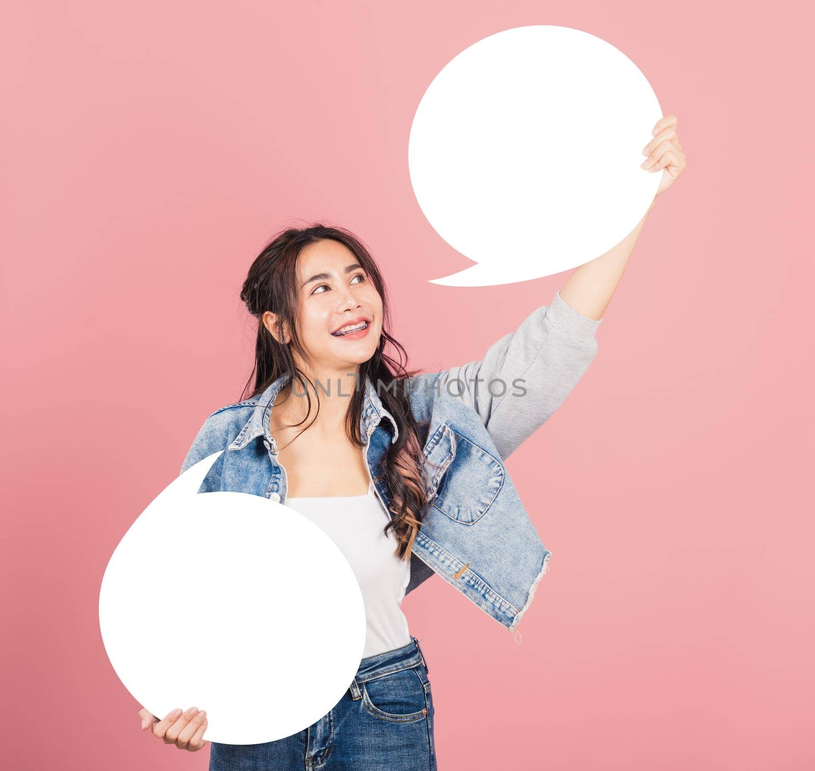 Happy Asian beautiful young woman smiling excited wear denims holding empty speech bubble sign, Portrait female posing show up for your idea looking at bubble, studio shot isolated on pink background