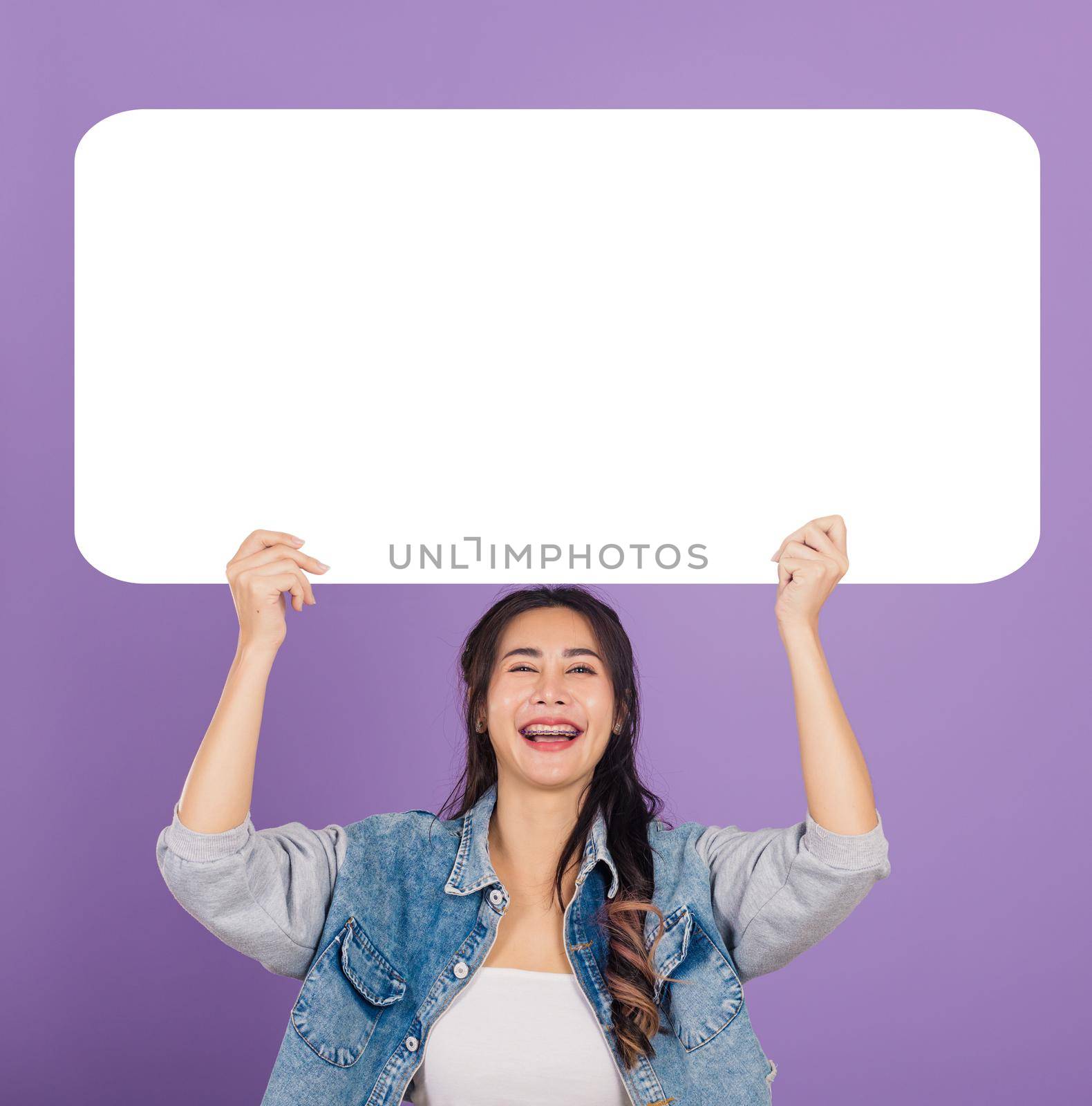 Happy Asian beautiful young woman smiling excited wear denims hold empty speech bubble sign, Portrait female posing show up for your idea looking at camera, studio shot isolated on purple background