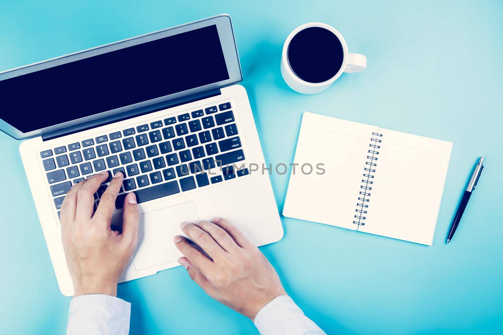 Hand of businessman working on laptop computer with notebook and cup of coffee on desk in office, hand of person typing keyboard with book and pen, top view, flat lay, business concept.