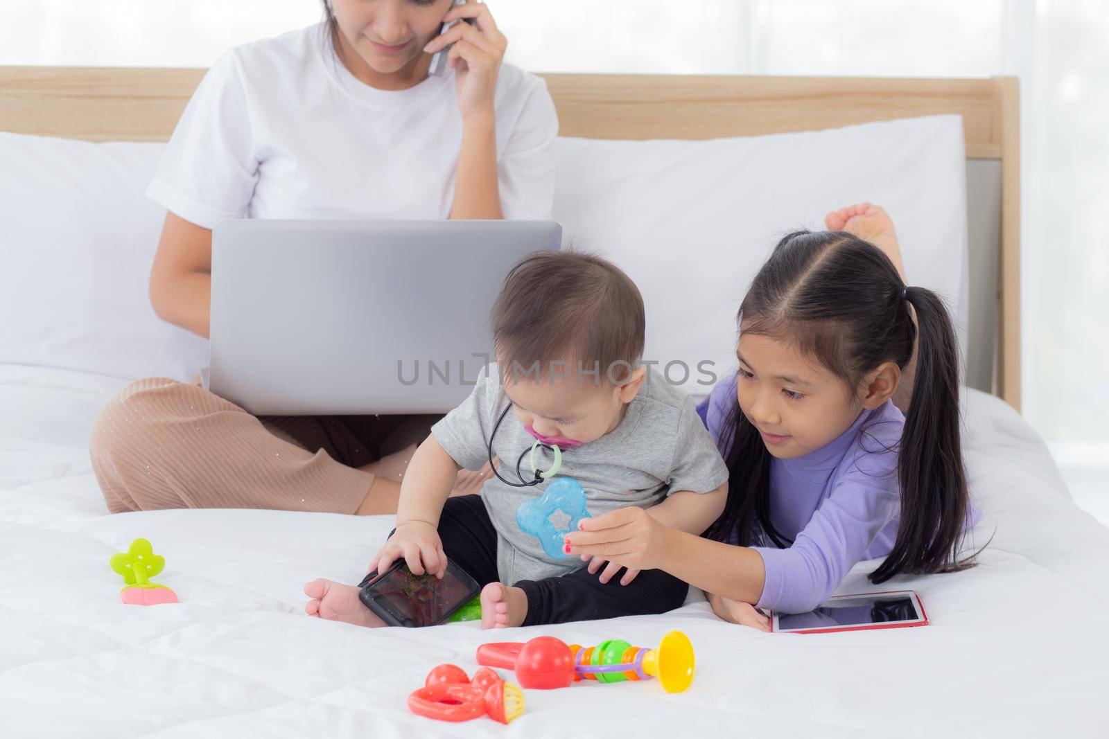 Young asian mother working at home online to internet with laptop computer and talking phone and daughter looking tablet on bed at bedroom, business woman is freelance, new normal, family concept. by nnudoo