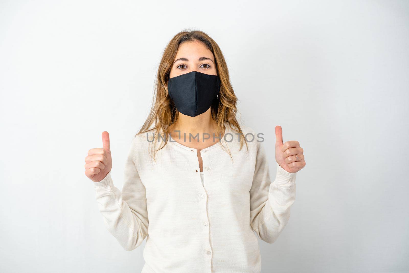Studio shot of Caucasian confident young woman show hands with thumbs-up gesture wearing Coronavirus protective mask on white background. Concept of thinking positive for the future due to Covid-19