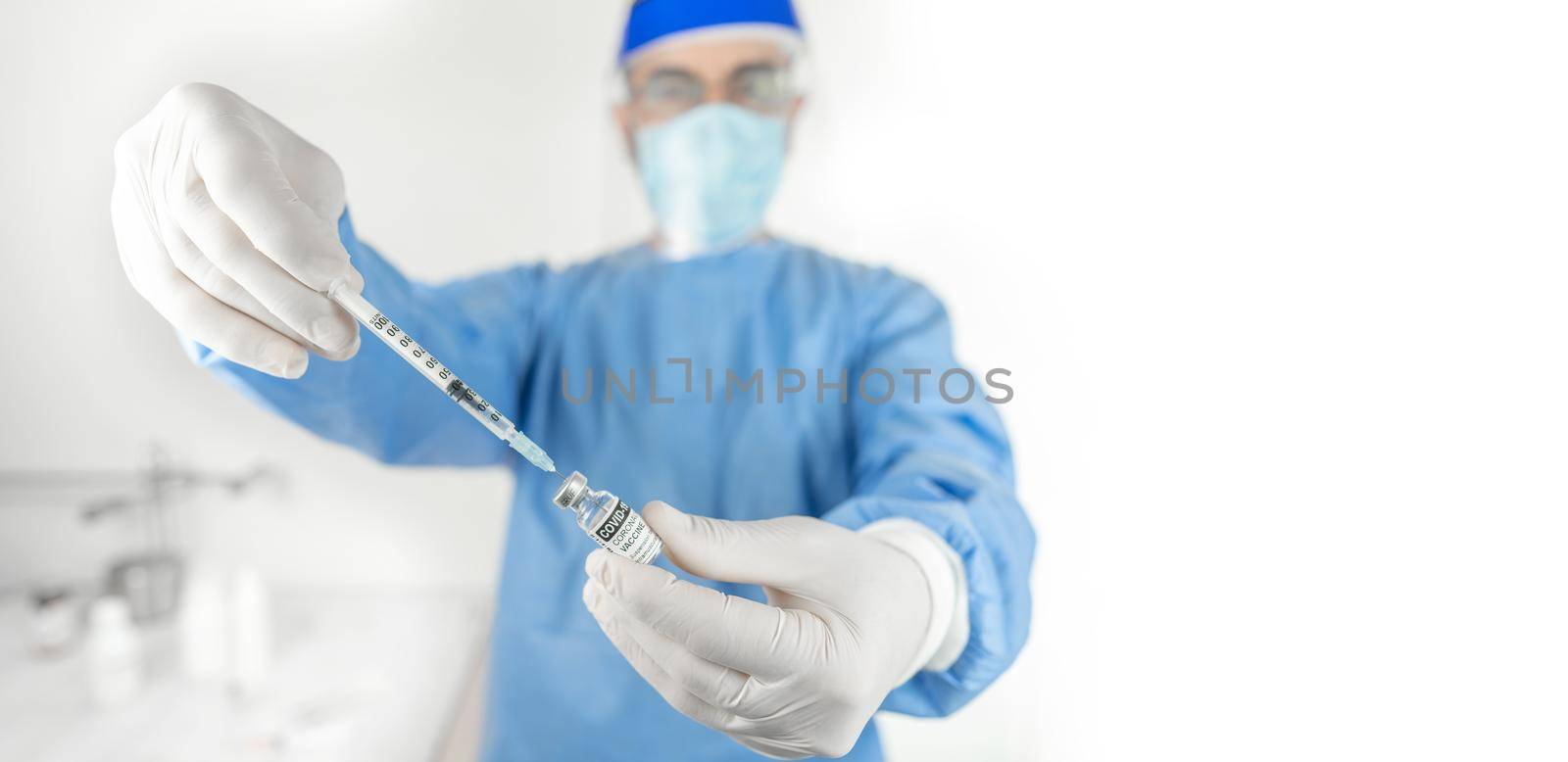 Senior doctor or healthcare worker in personal protective kit in hospital or health clinic interior preparing syringe for a shot of drug as treatment amid Coronavirus or COVID 19 epidemic or pandemic by robbyfontanesi