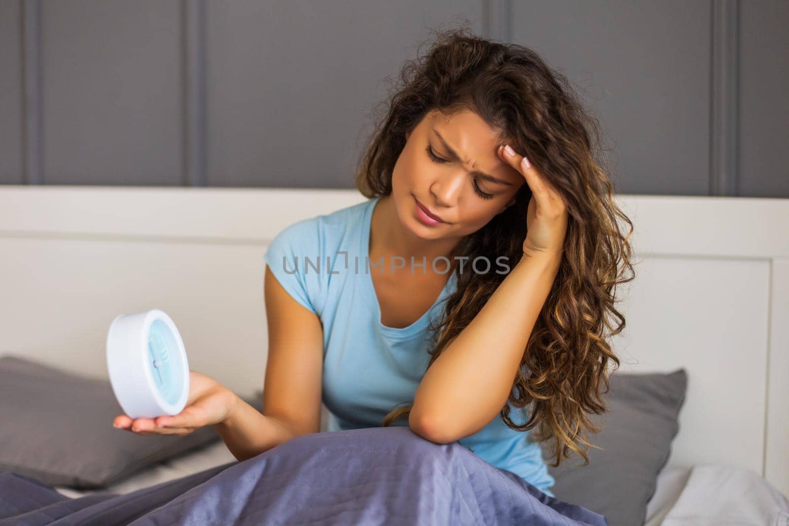 Sad woman in pajamas holding alarm clock in bed.