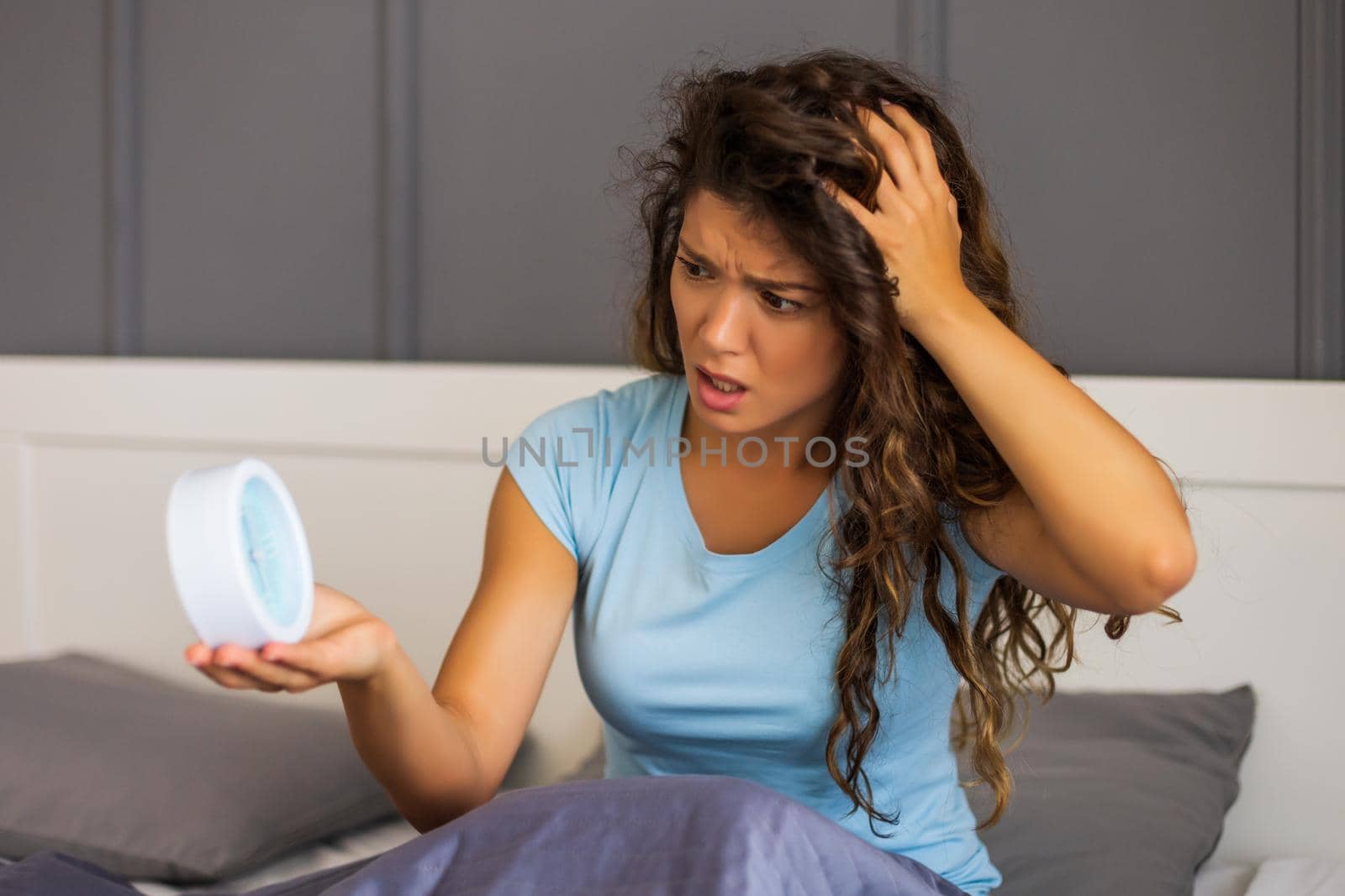 Angry woman in pajamas holding alarm clock in bed.