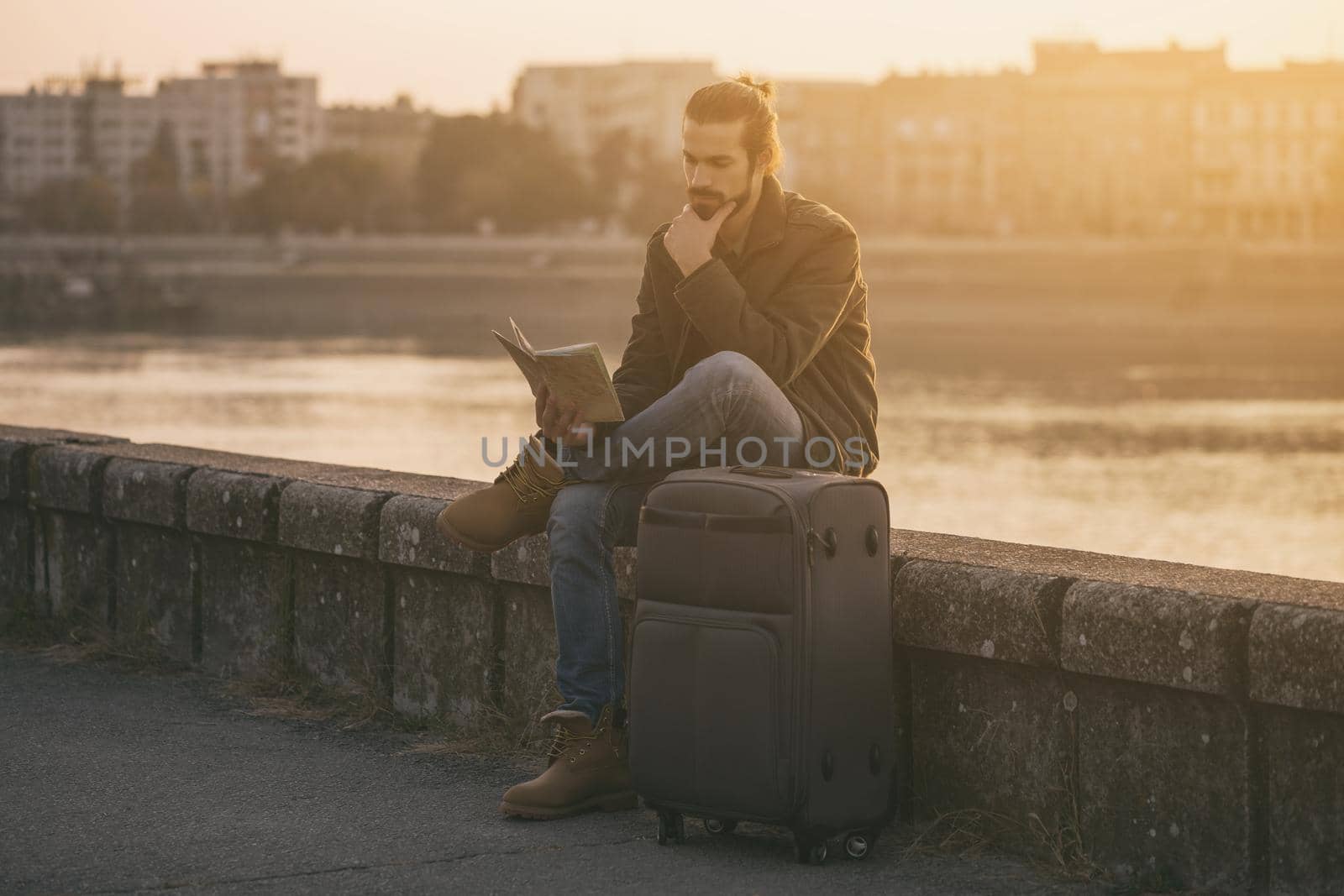 Confused businessman with suitcase and city map sitting lost by the river.