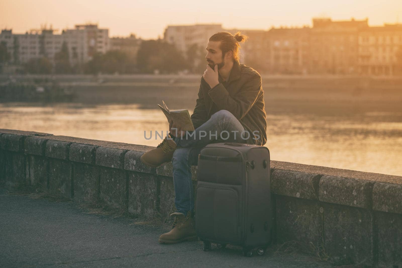 Confused businessman with suitcase and city map sitting lost by the river.