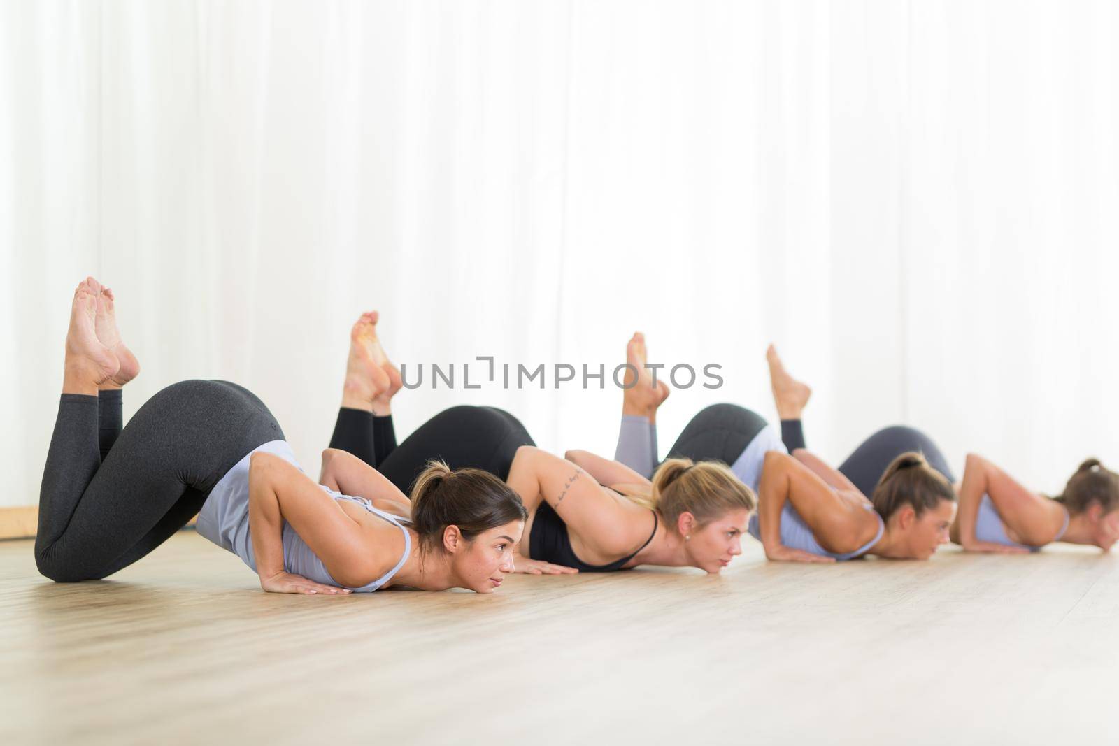 Group of young sporty sexy women in yoga studio, practicing yoga lesson with instructor, forming a line in Shishosana bent puppy dog asana pose. Healthy active lifestyle, working out indoors in gym.