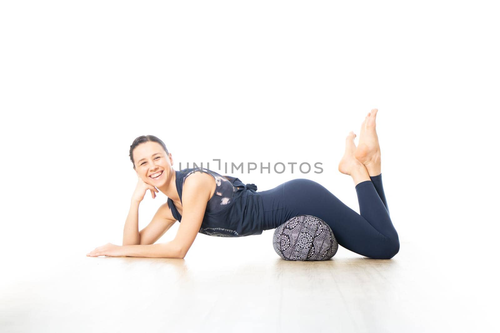 Restorative yoga with a bolster. Young sporty female yoga instructor in bright white yoga studio, lying on bolster cushion, stretching, smilling, showing love and passion for restorative yoga by kasto