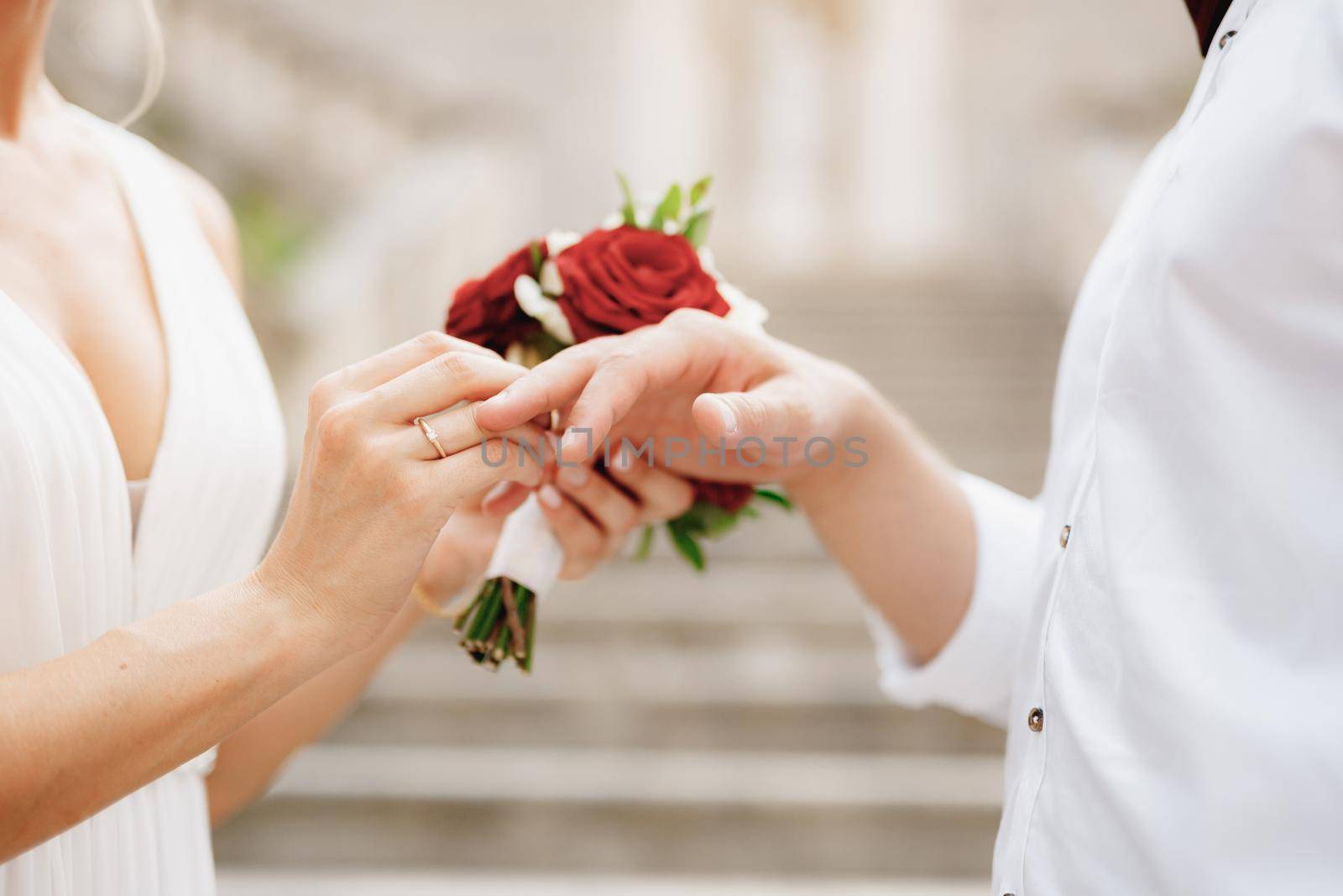The bride puts a wedding ring on the groom's finger and holds a bouquet of red and white roses in her hand . High quality photo