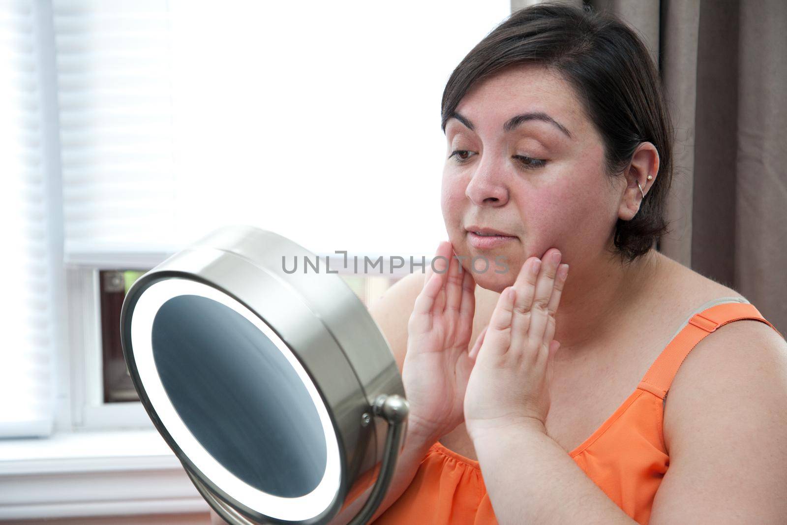 Sitting beside a window with a vanity mirror, a woman touches her face before applying makeup.