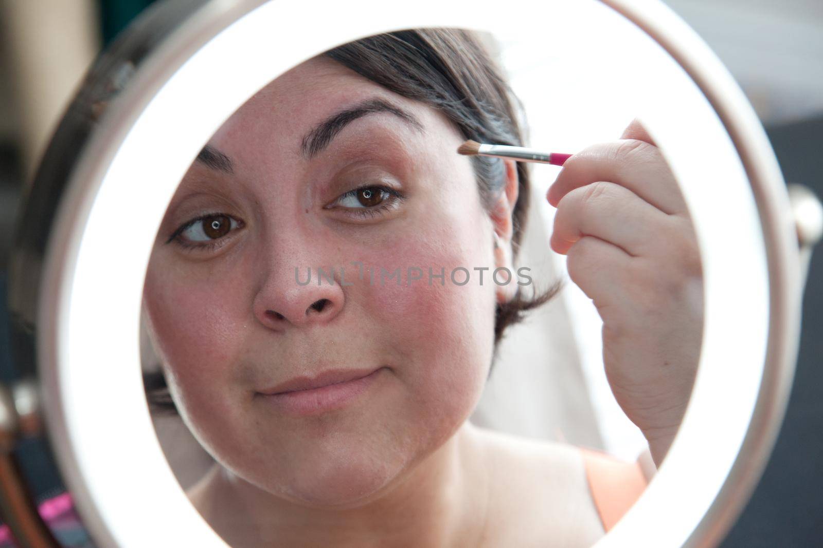 Looking in a round vanity mirror, a woman holds a brush to apply makeup 