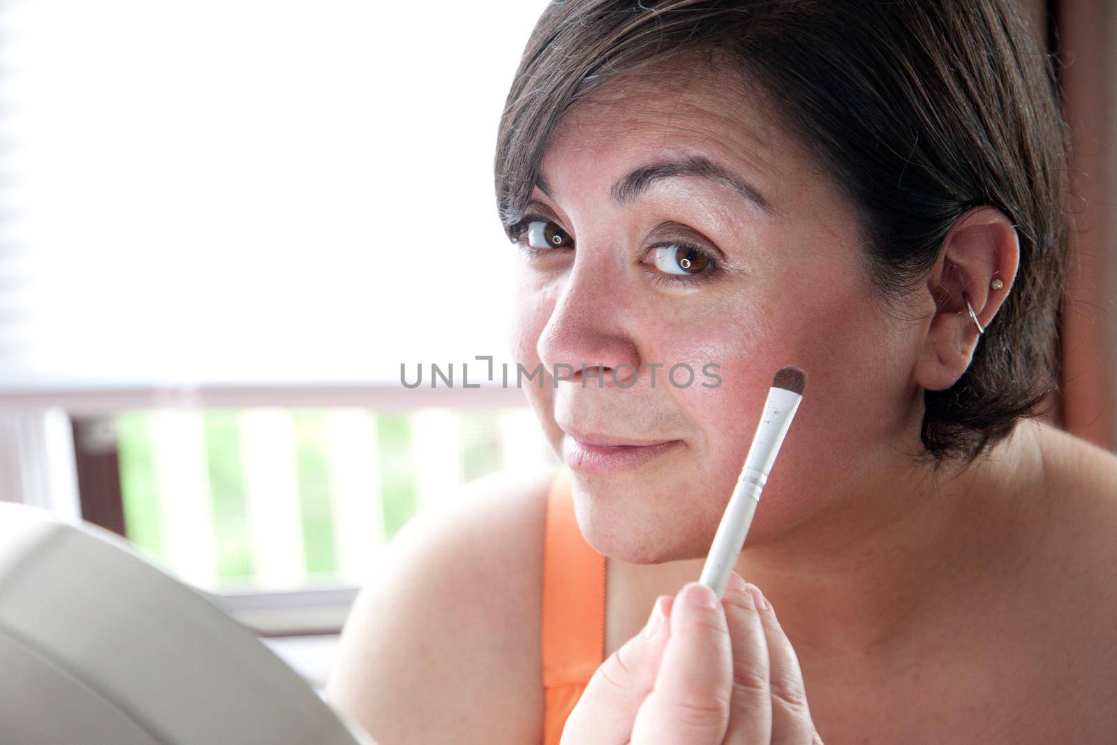  A dark haired latina woman smiles at you as she applies her makeup by a mirror 