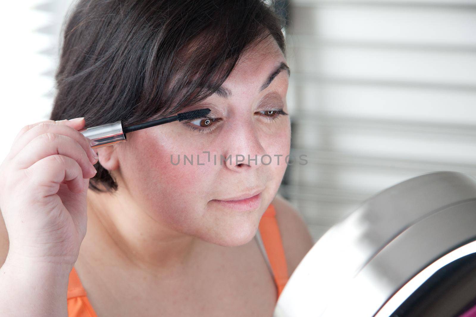 Woman in front of mirror puts holds a mascara wand and applies eye makeup 