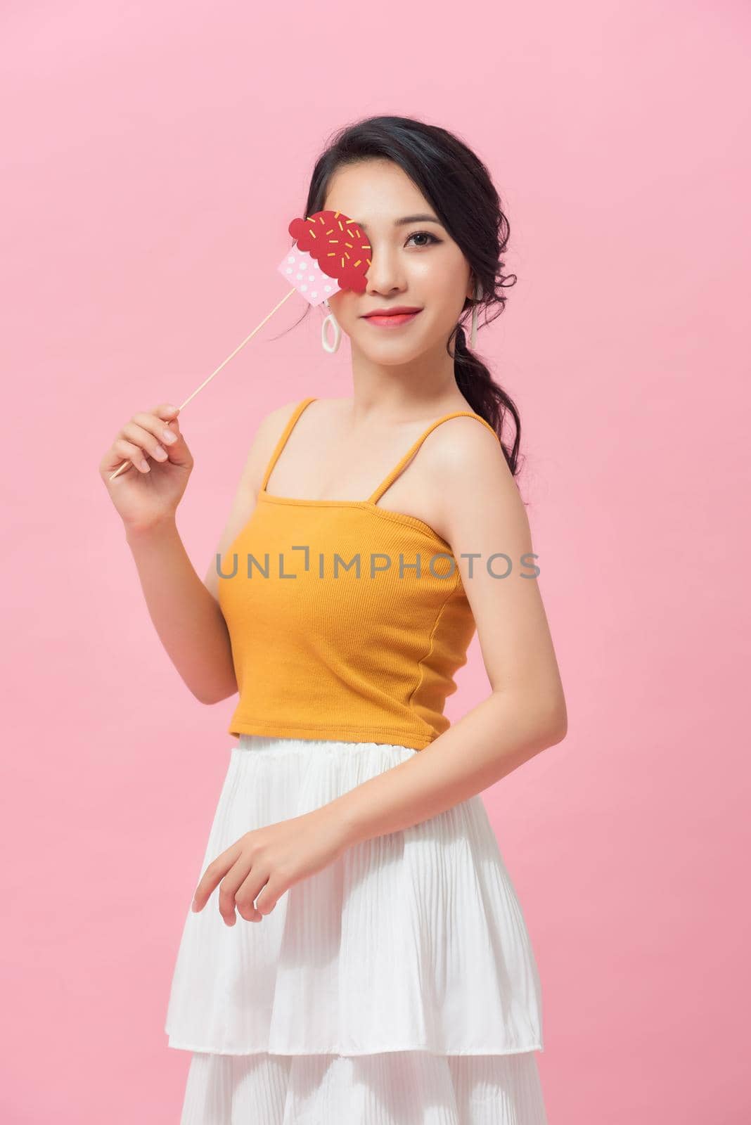 Happy young woman holding paper cupcake on stick over pink background